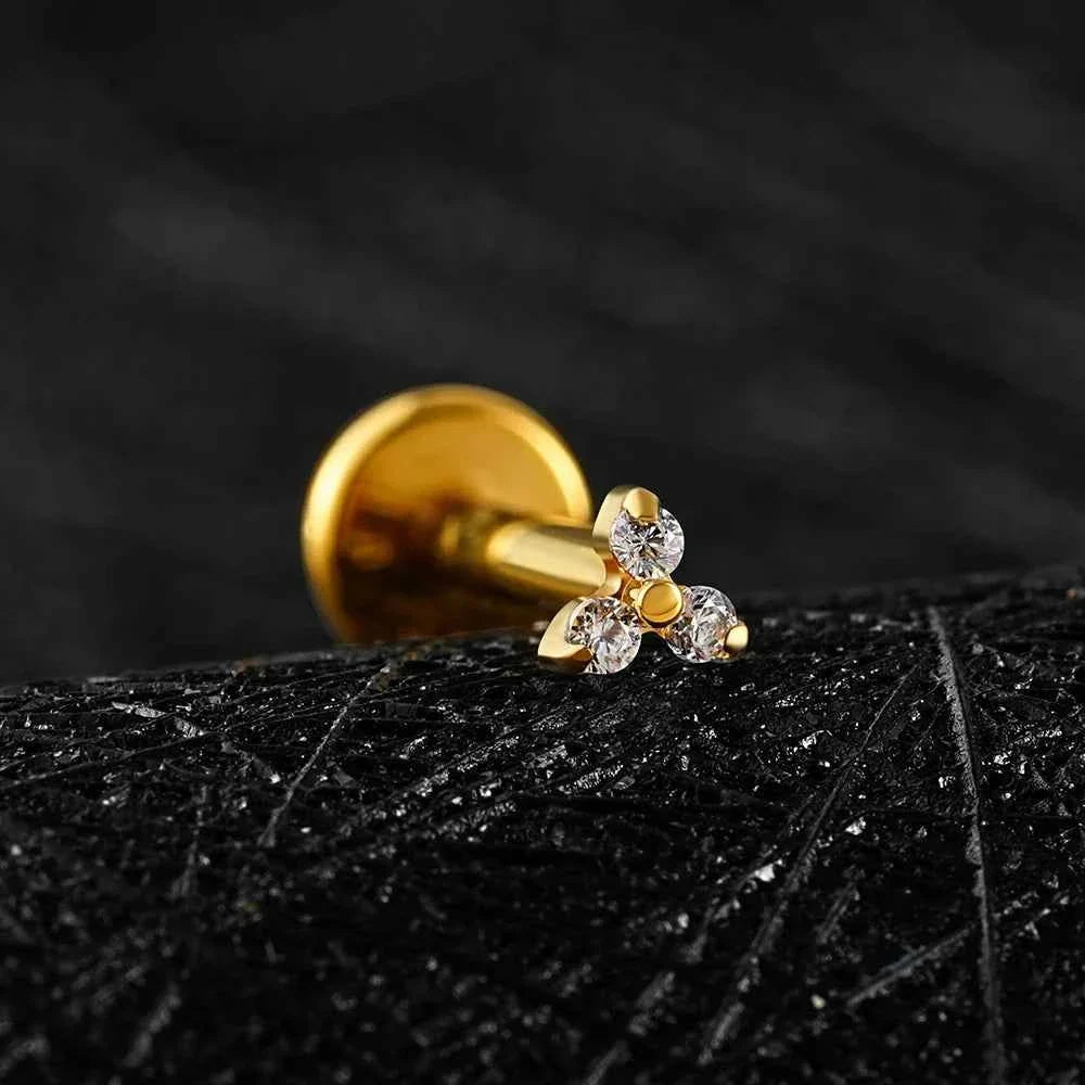 Gold-tone Trinity CZ flat back stud resting on dark textured stone.